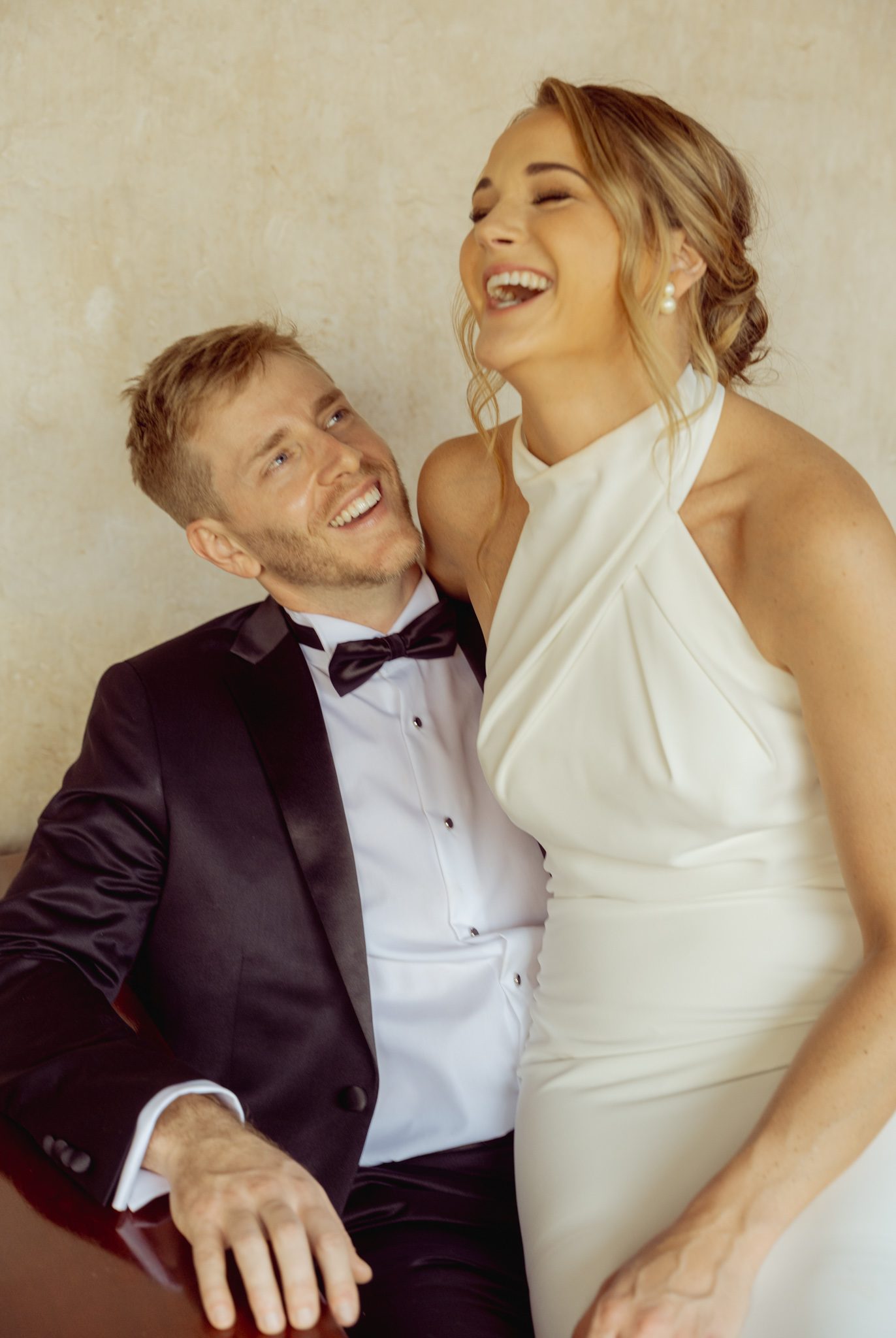 Bride and groom laughing together against a pale plaster wall