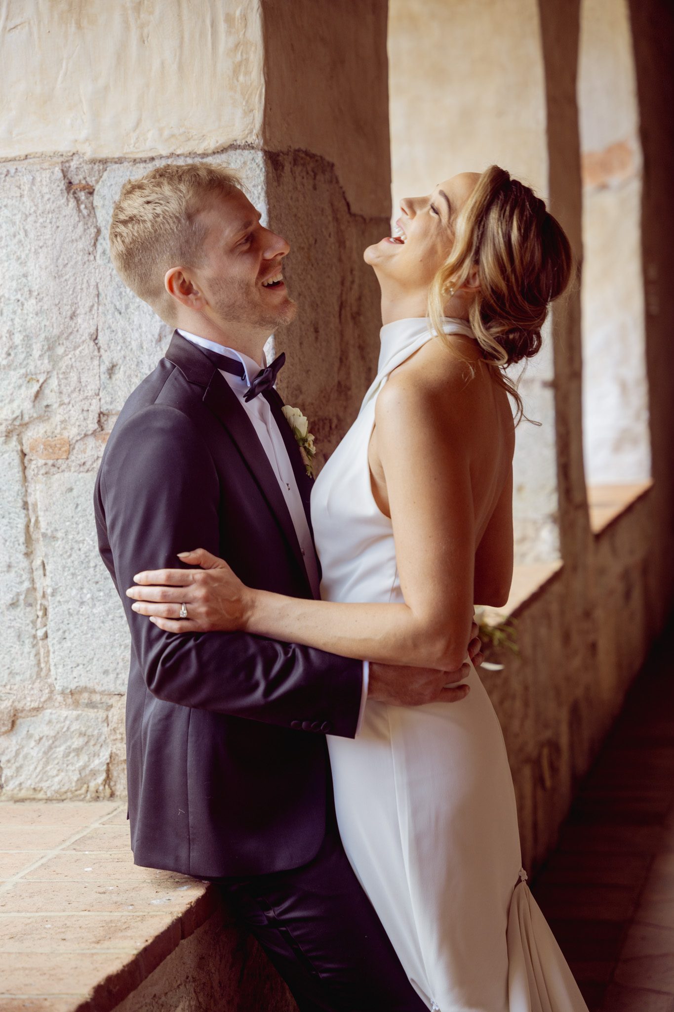 Bride laughing with her head tipped back as the groom holds her beside a stone window