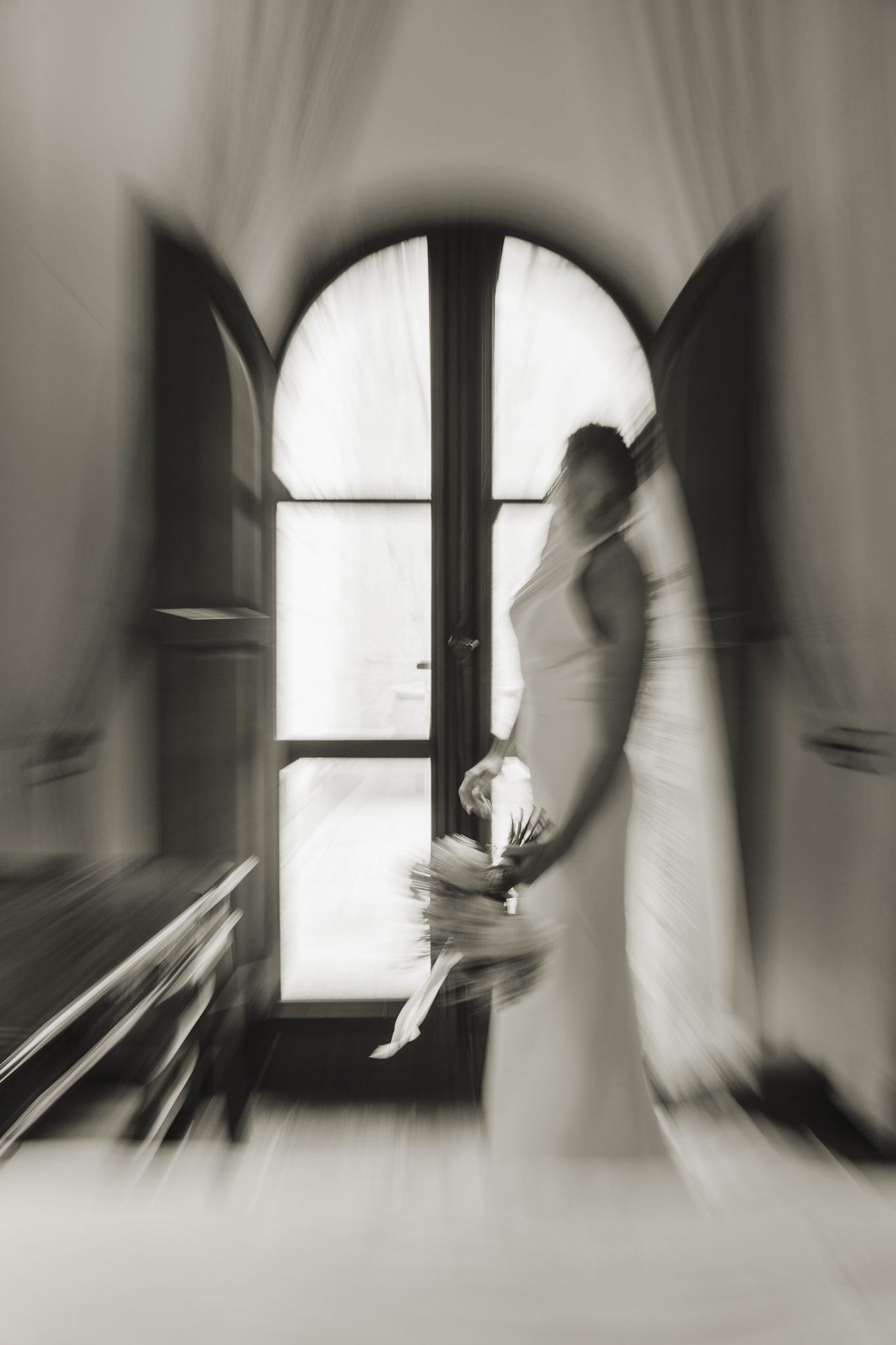 Black and white photo of a bride standing at a tall arched window, the room blurred around her