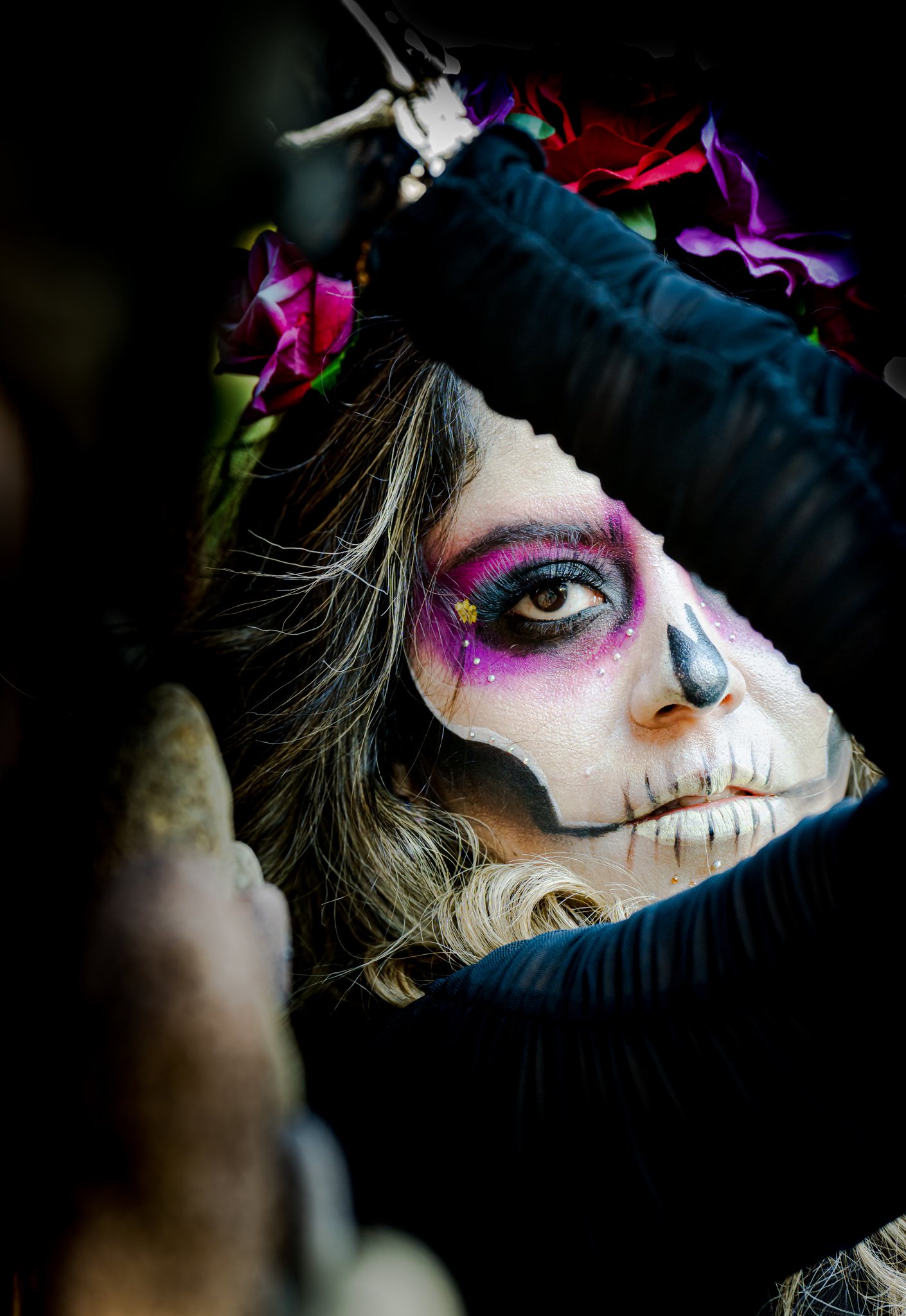 A catrina with purple eye paint framed by her raised arm and a crown of flowers