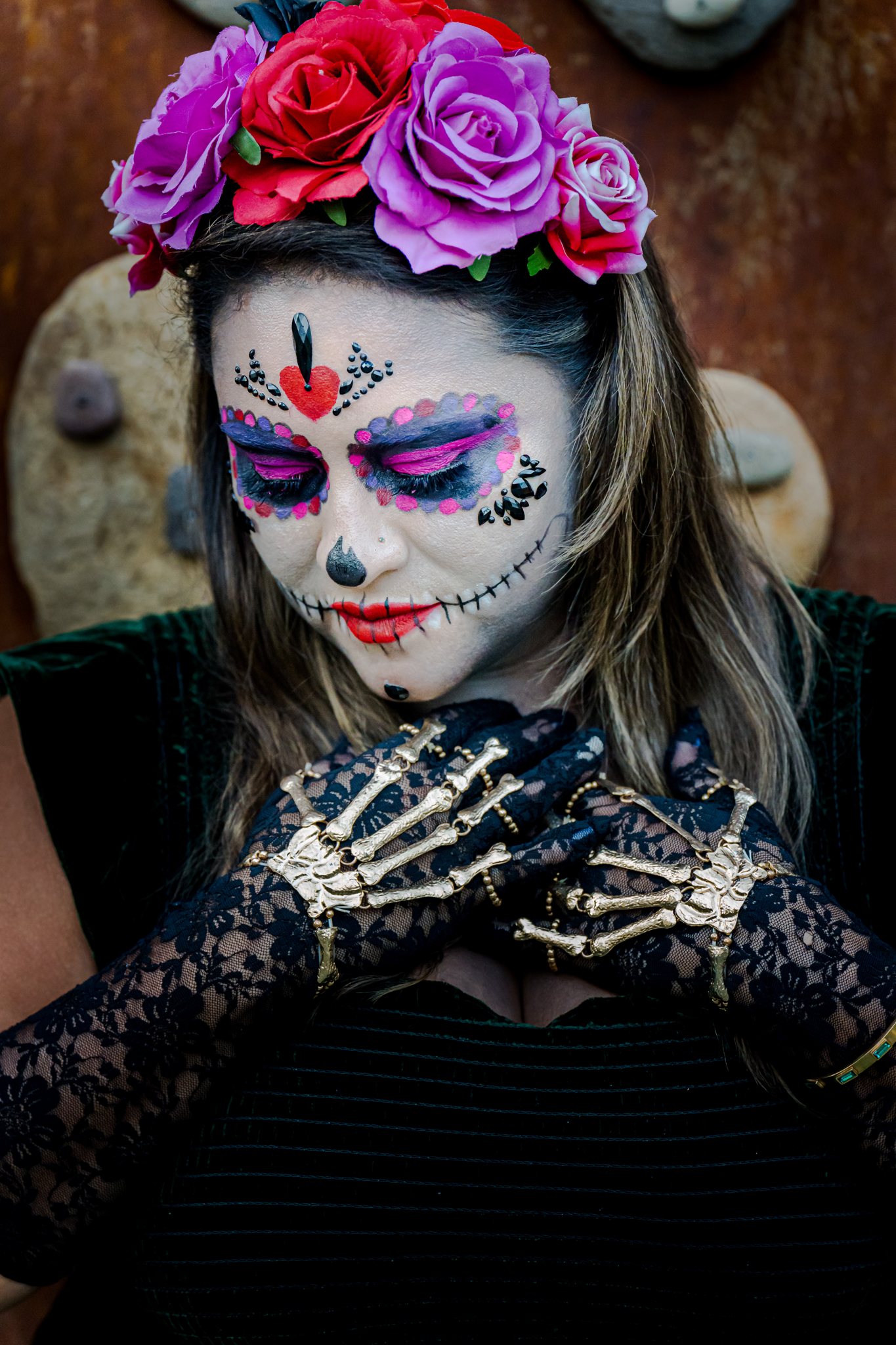 A catrina in a crown of roses looking down, her hands crossed in skeleton lace gloves