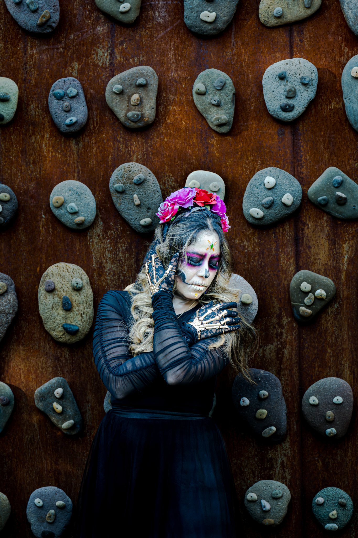 A catrina in skeleton gloves against a rust wall set with carved stone faces