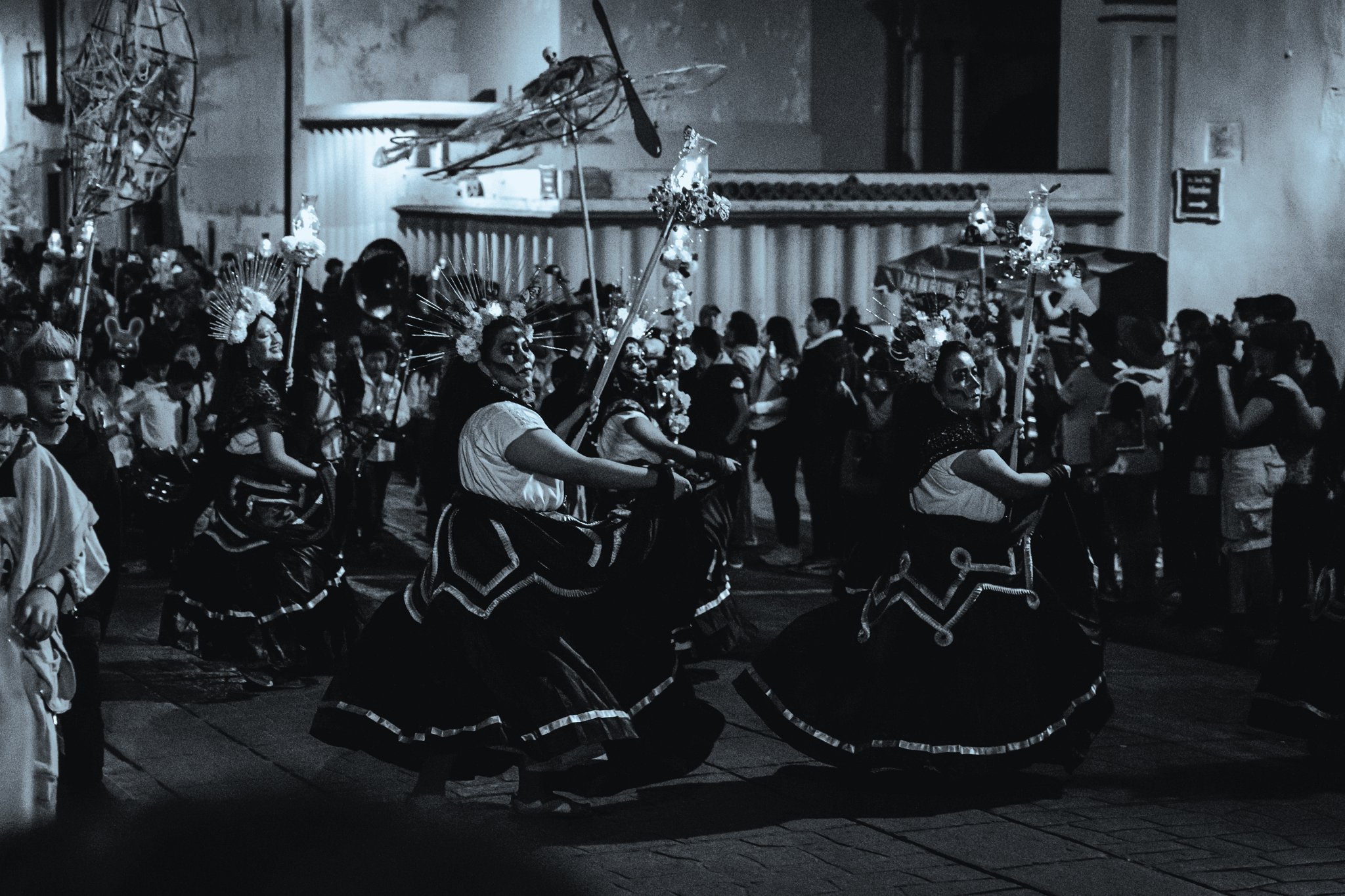 Catrinas in wide black skirts and lit headdresses dancing in a Día de Muertos comparsa through the streets of Oaxaca at night