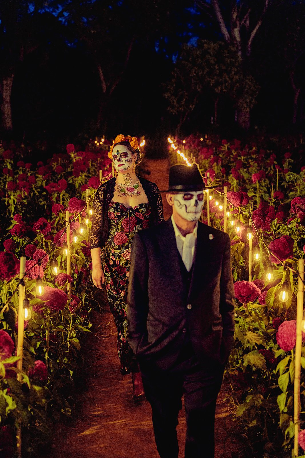 A catrín and catrina at night on a path of candles through cempasúchil and cockscomb