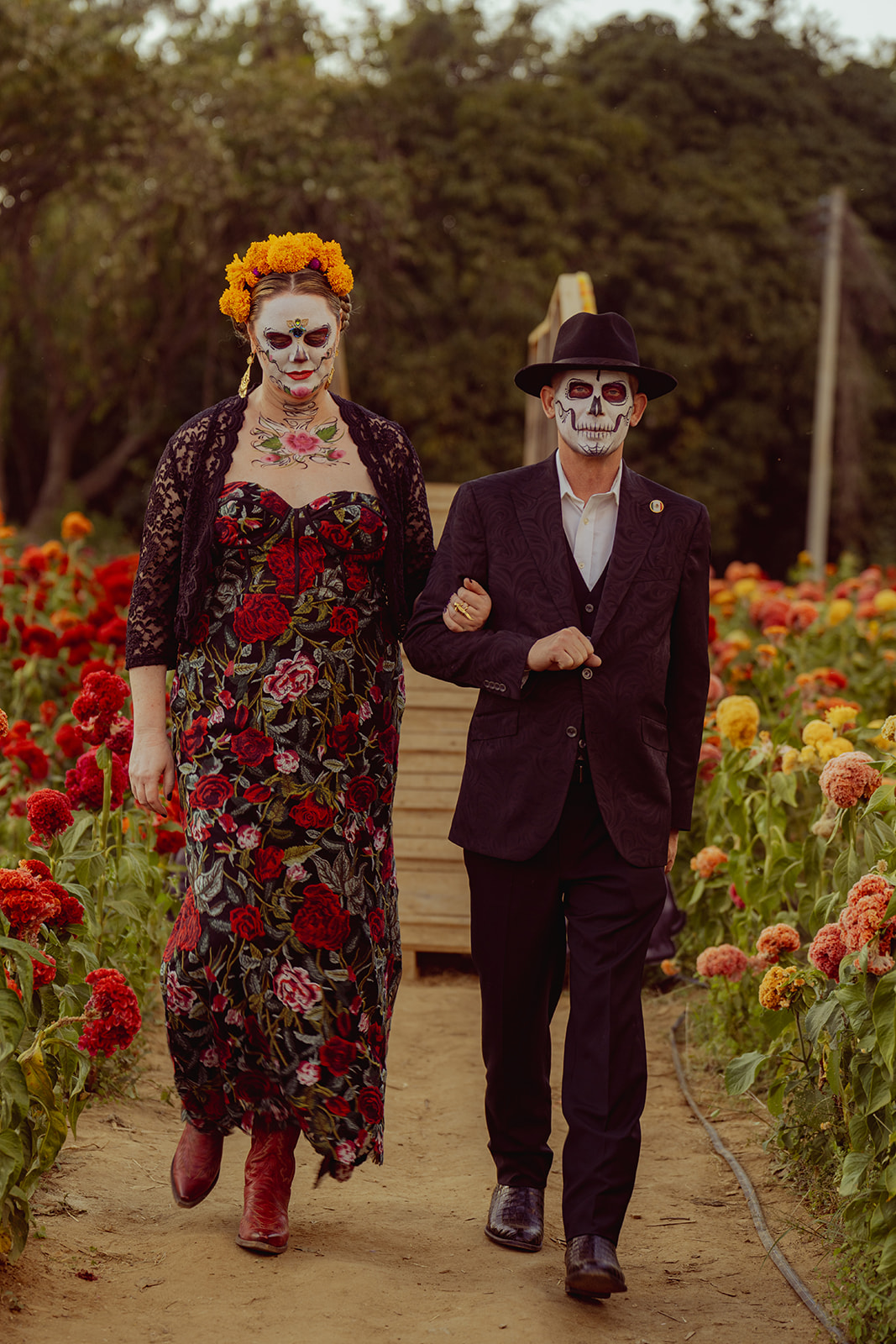 A catrina and catrín walking arm in arm through a field of cempasúchil and cockscomb
