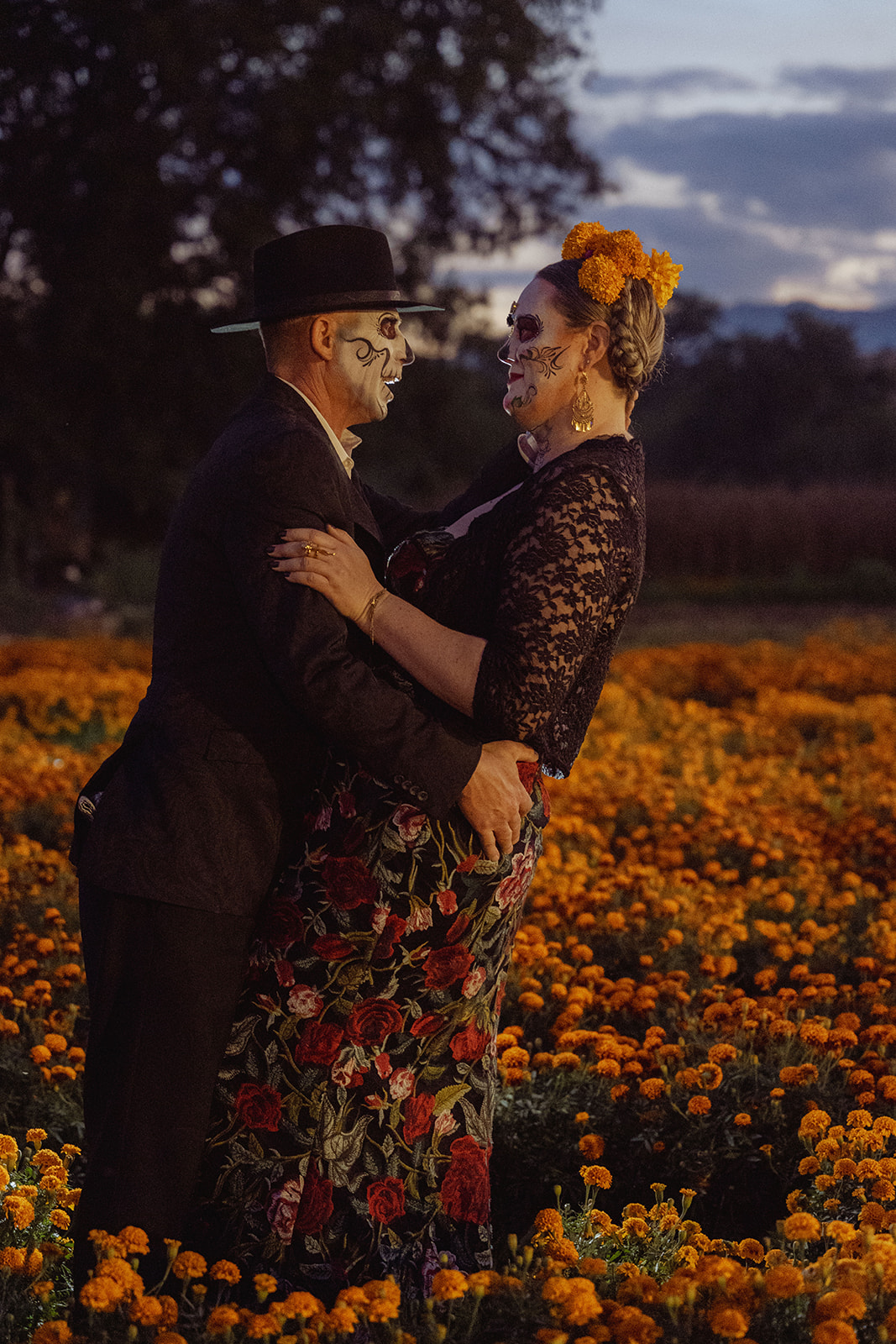 A catrina and catrín holding each other in a marigold field at dusk
