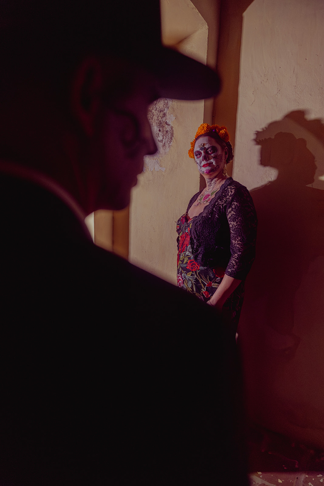 A catrín in shadow in the foreground and a catrina lit against a plain wall behind him