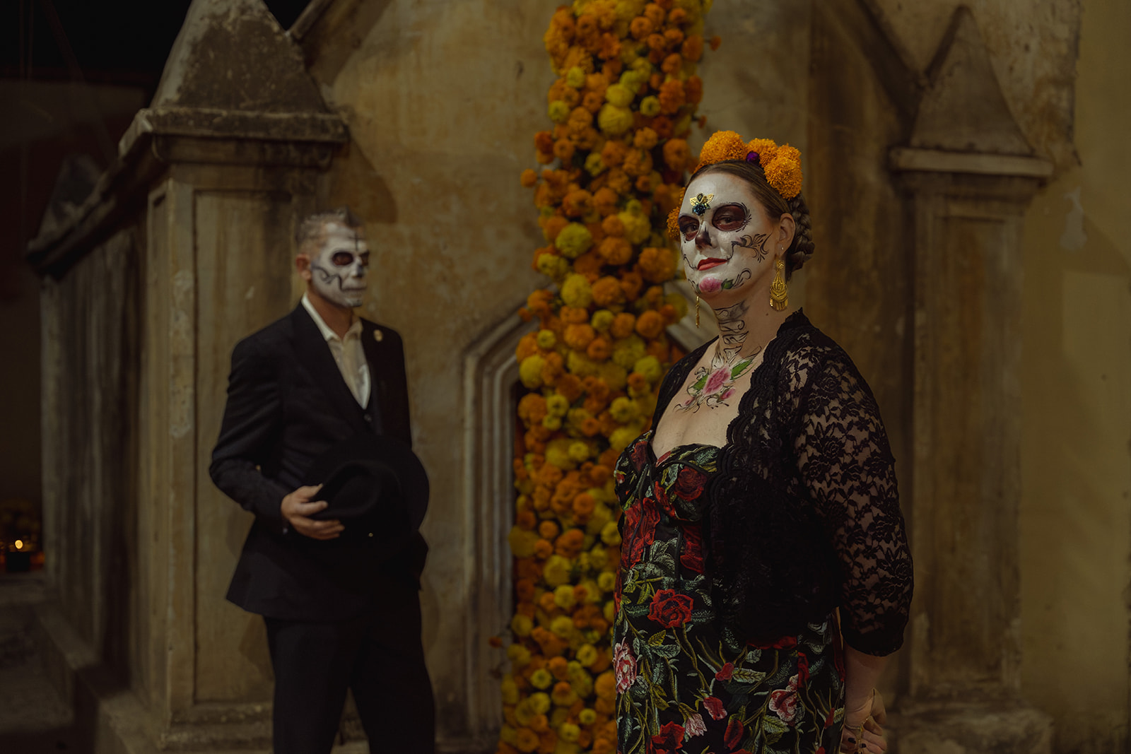 A catrina and catrín side by side at night beside a column of marigolds
