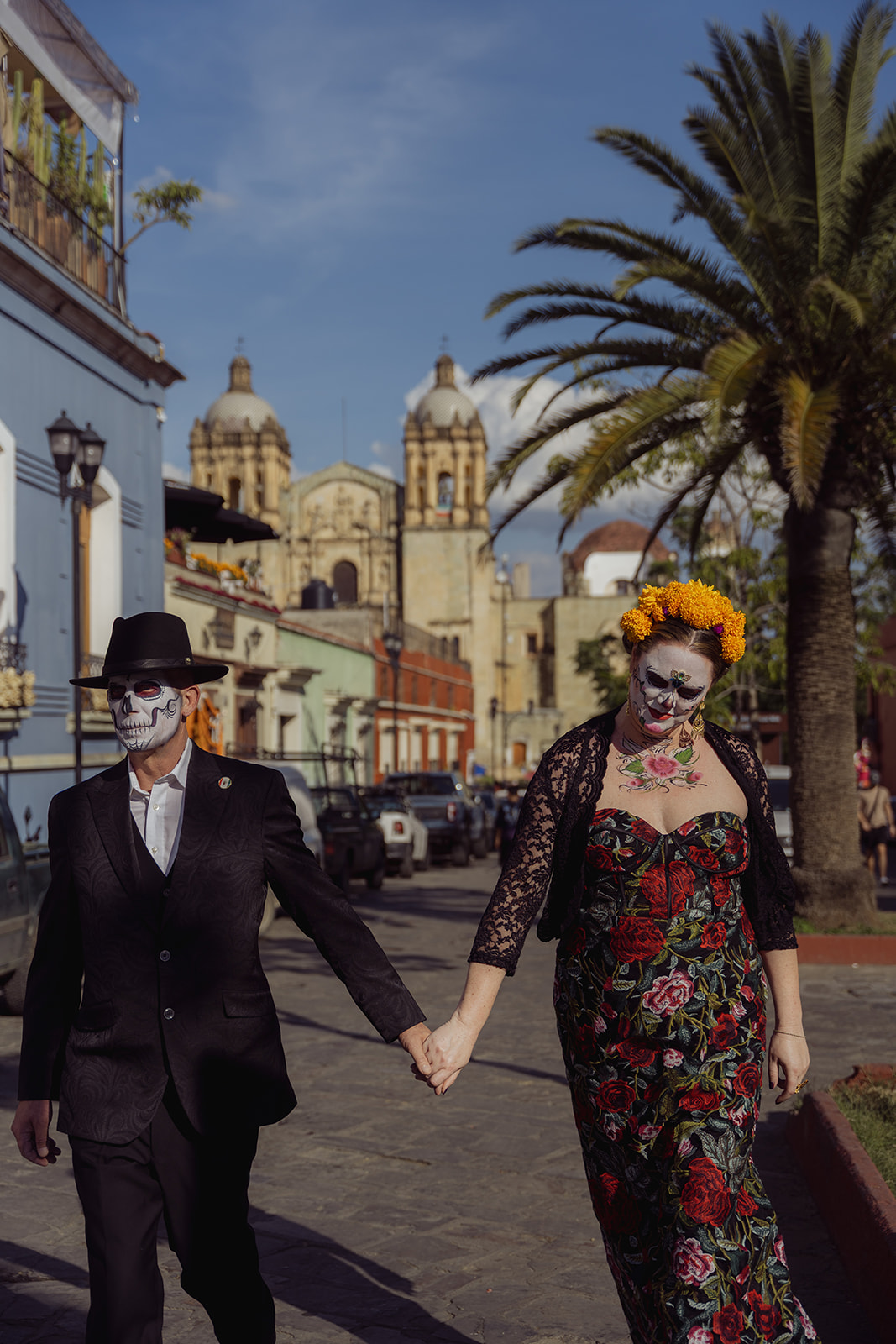A catrina and catrín walking hand in hand down Calle Alcalá with Santo Domingo behind them