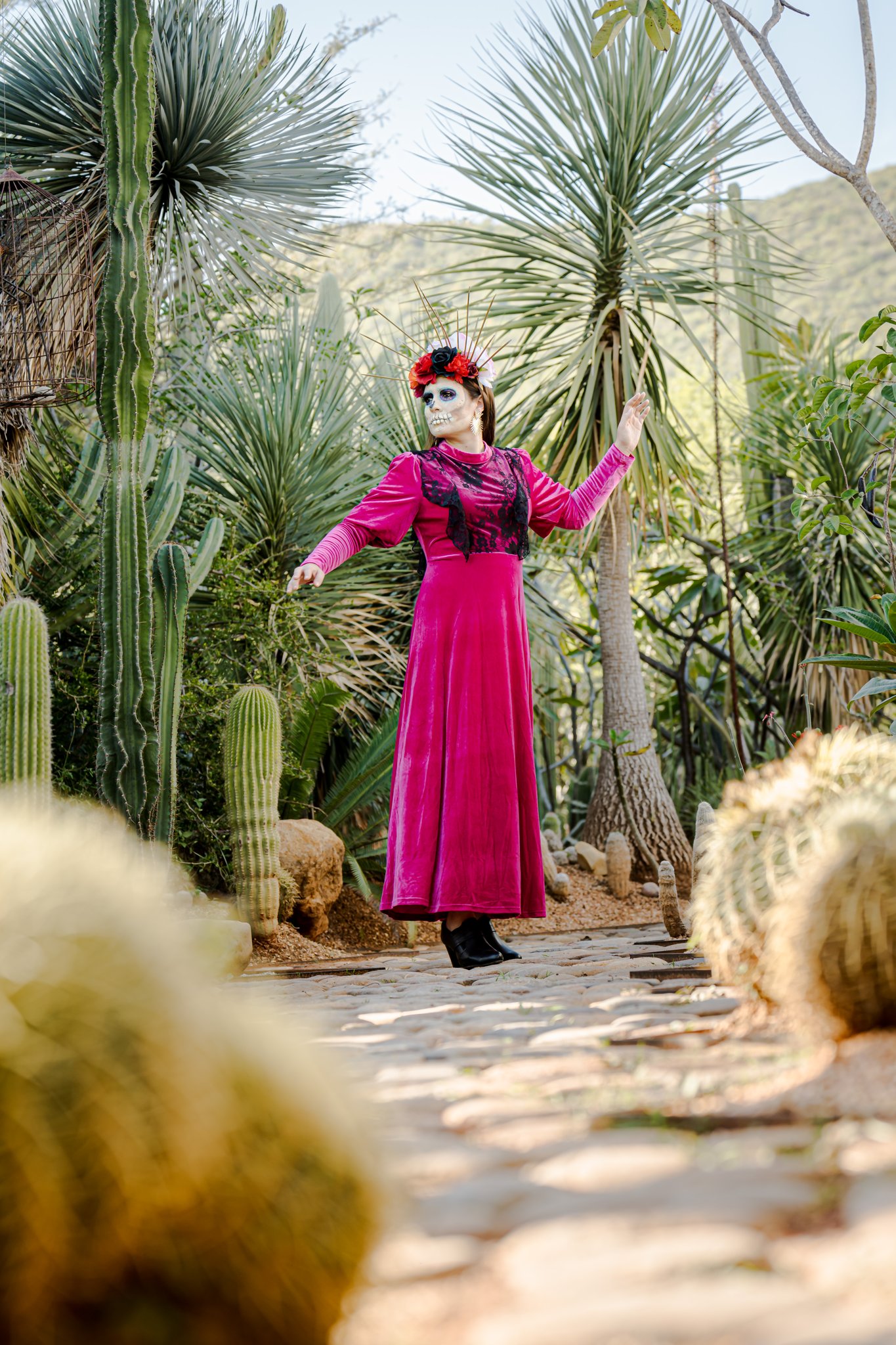 A catrina in a long magenta dress standing on a stone path among tall cactus and palms at Vives Verde