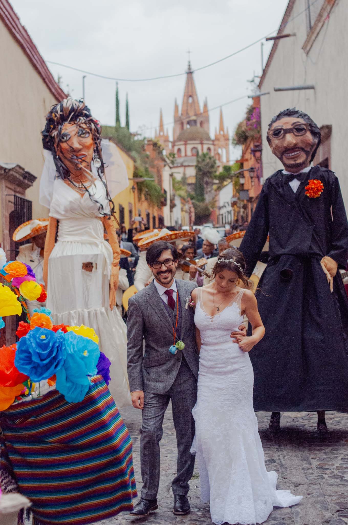 Bride and groom walking a cobblestone street with giant mojigangas, the Parroquia behind them