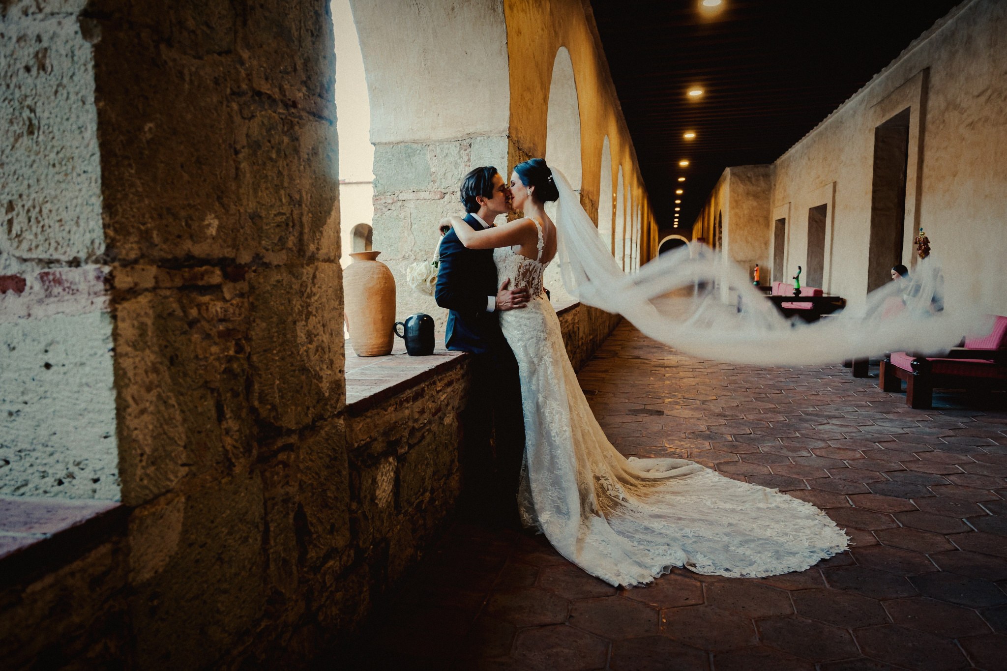 A couple kissing in a hacienda corridor as her veil streams out behind them, Oaxaca