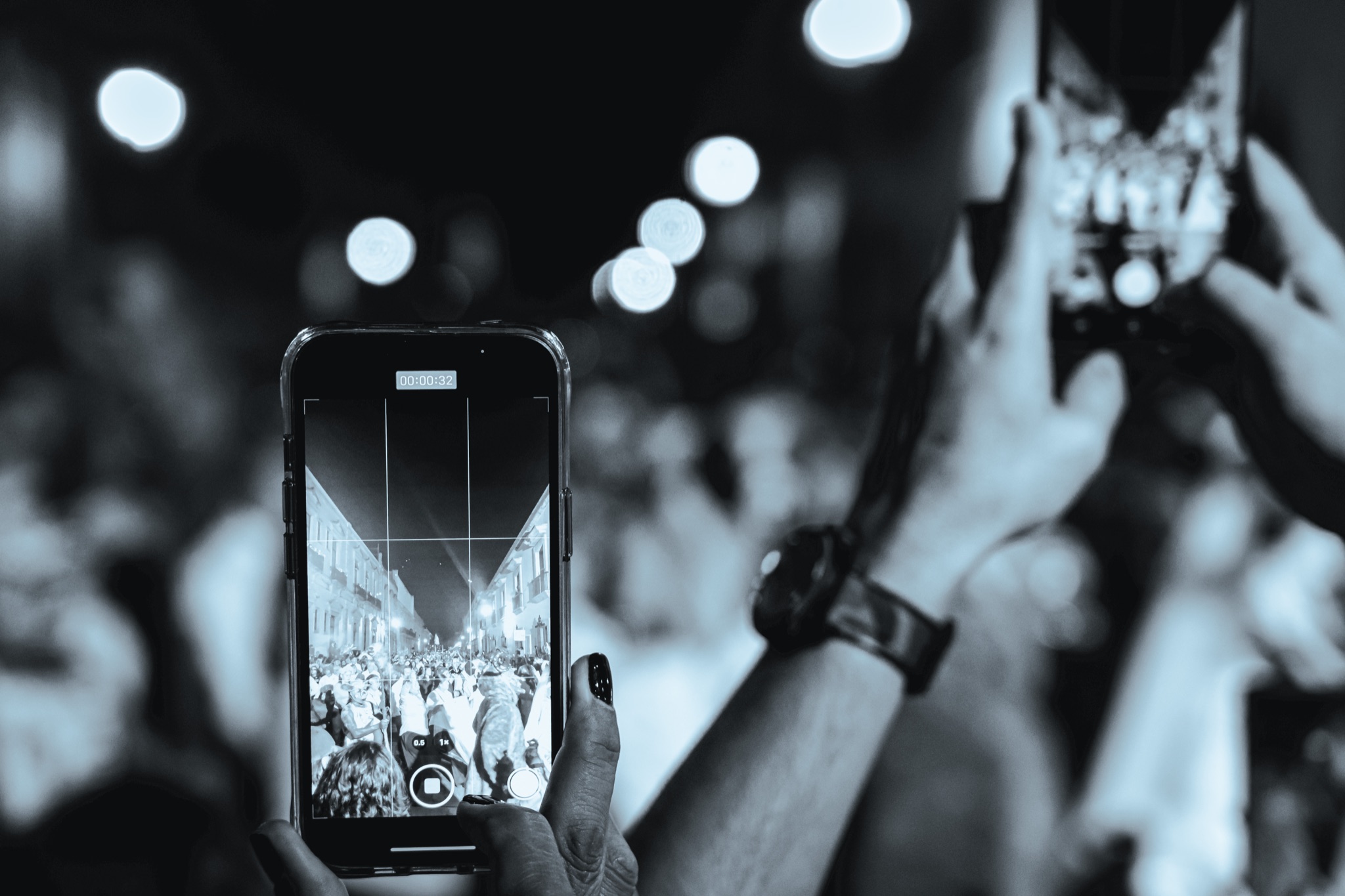 A hand holding a phone, filming a Día de Muertos comparsa at night, in black and white