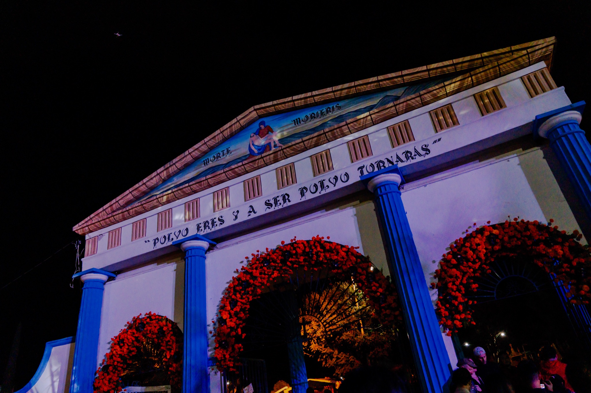 A cemetery entrance at night, with blue columns and arches of red flowers